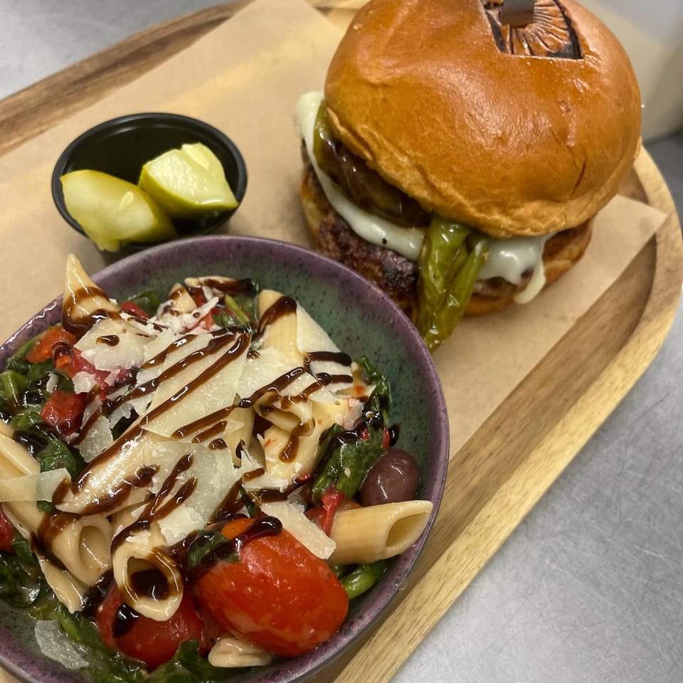 A burger with melted cheese and green peppers, pasta salad, and pickles on a wooden tray.