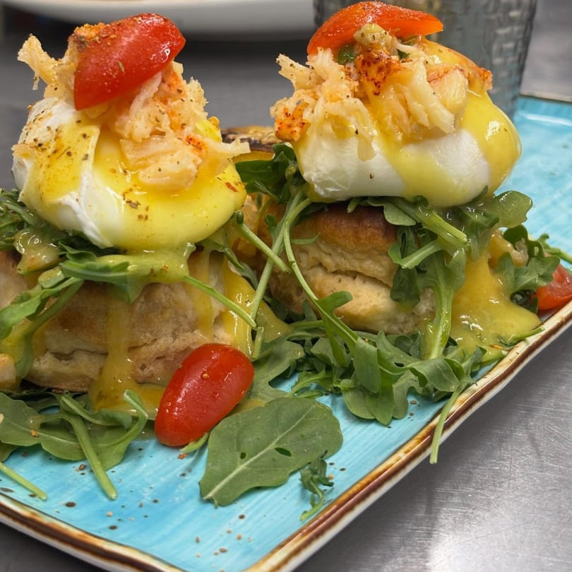 Two lobster eggs benedict with hollandaise sauce, arugula, and cherry tomatoes on a blue plate.