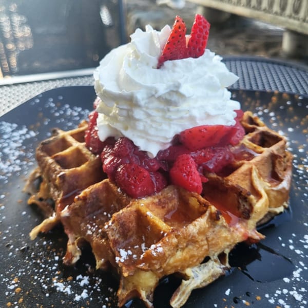 Golden waffle topped with whipped cream, fresh strawberries, and syrup on a black plate.