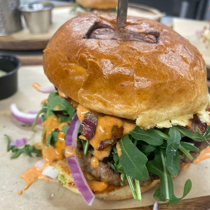 A Brunch House 1850 burger with arugula, red onion, bacon, and sauce, held by a knife.
