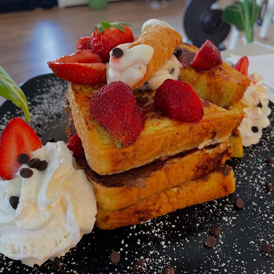 Stacked French toast with strawberries, whipped cream, chocolate chips, and a cannoli on a black plate.