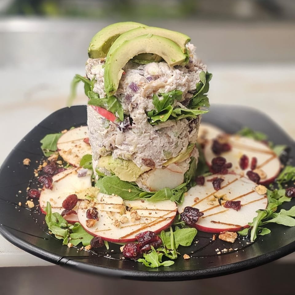 A tall chicken salad stack with avocado, arugula, and apple slices on a black plate.