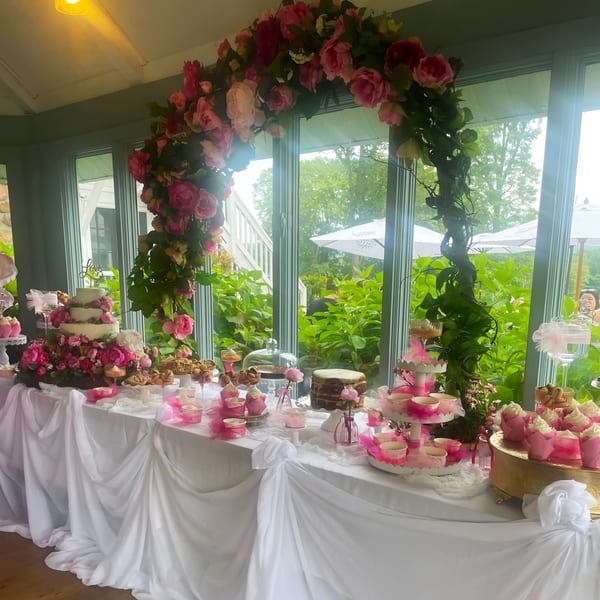A dessert table with cakes, cupcakes, and cookies, adorned with pink flowers and white draping.