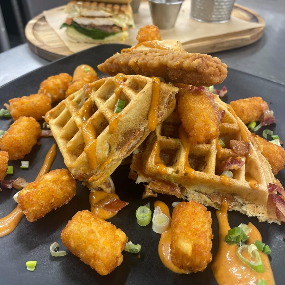 Chicken and waffles with tater tots, bacon bits, and sauce, with a sandwich in the background.