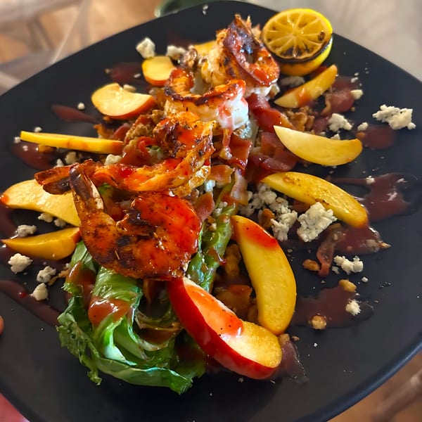 Grilled shrimp salad with peaches, blue cheese, and a lemon wedge on a black plate.