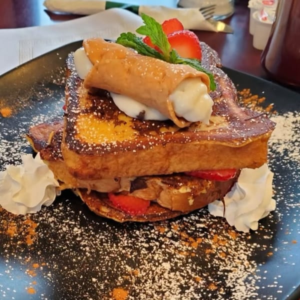 Cannoli-topped French toast with strawberries, whipped cream, and powdered sugar on a black plate.
