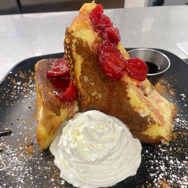 Stuffed French toast with strawberries, whipped cream, powdered sugar, and syrup on a black plate.