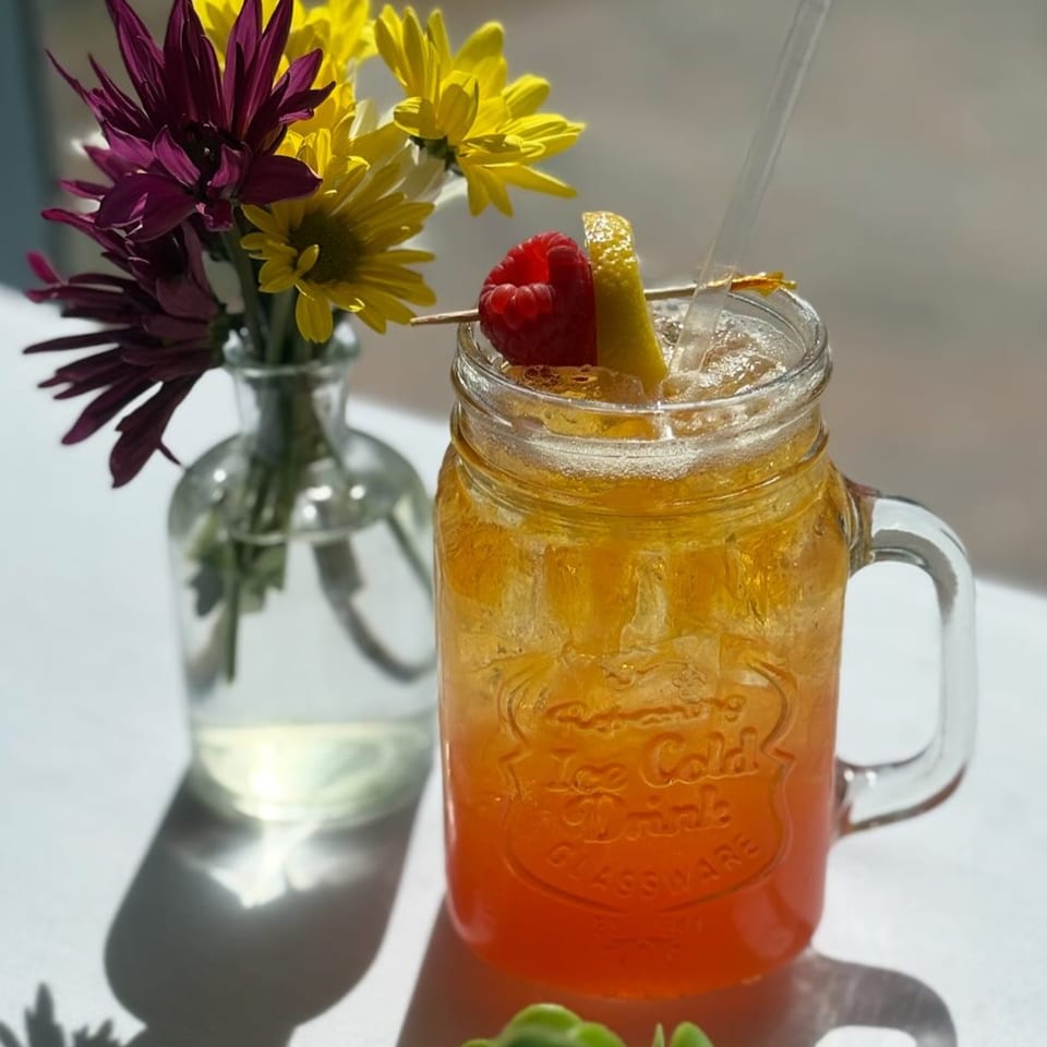 A refreshing iced drink with raspberry and lemon garnish next to a vase of purple and yellow flowers.