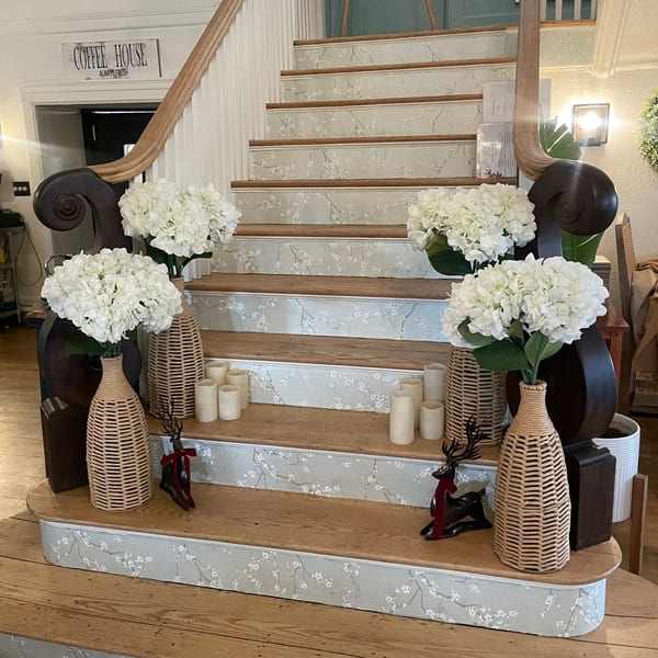 A decorative staircase with floral wallpaper risers, white hydrangeas in wicker vases, and candles.